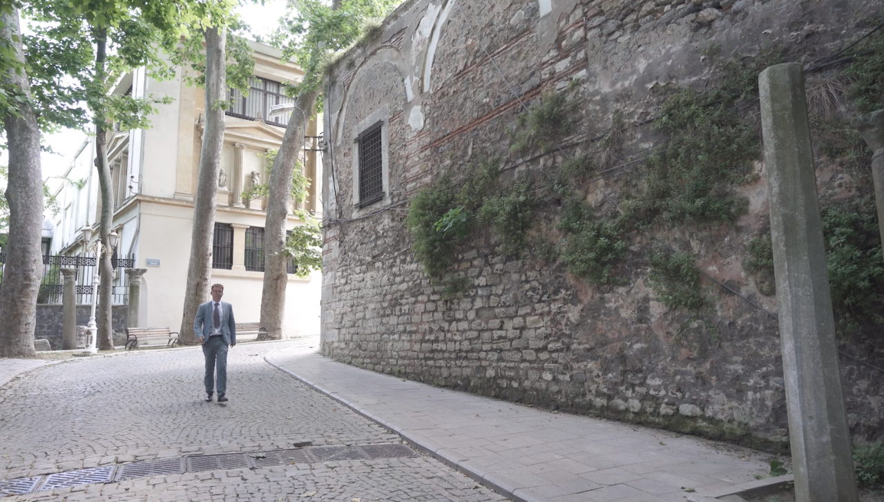 Dr Mansfield walks past a very old stone wall on a cobblestone street